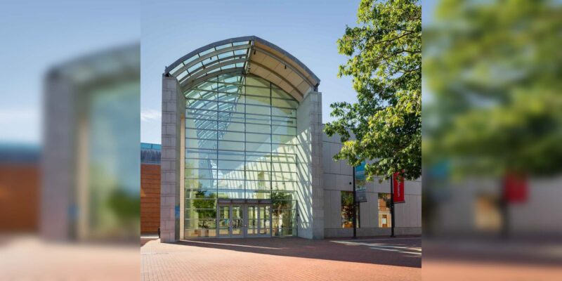 The exterior entrance of a museum featuring a tall, arched glass facade and a brick plaza.