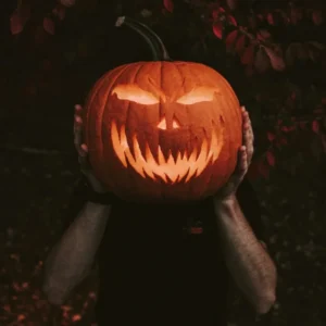 A person holds a glowing, carved jack-o’-lantern in front of their face at a Sevenings event.