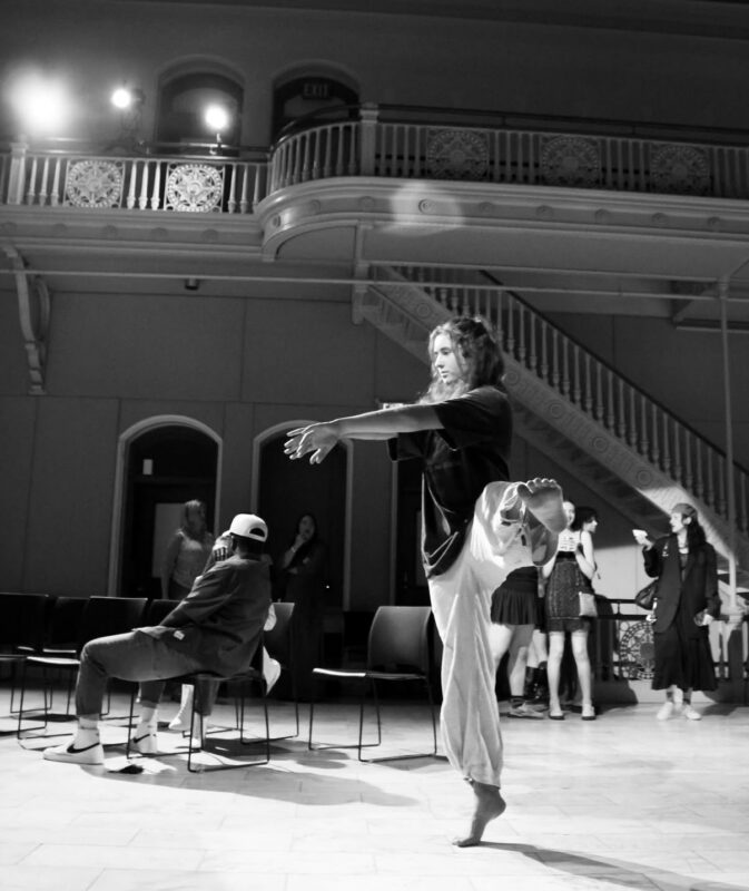 A person practices a dance class pose, stretching on one leg with arms extended and holding their other foot. Others watch in a spacious room with a staircase and ornate balcony, evoking the spirit of contact improvisation. The image is in black and white.