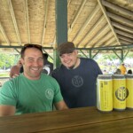 Two men standing at a table with cans of beer.