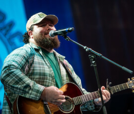 A bearded guitarist performs beneath a large Patrick's Pack logo projected on stage at A Night for Patrick in Beverly