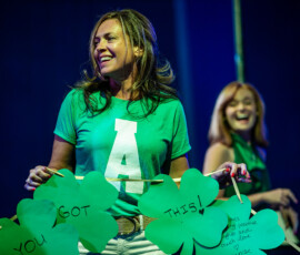A volunteer in a green Patrick's Pack shirt holds up oversized paper shamrocks reading You Got This at A Night for Patrick