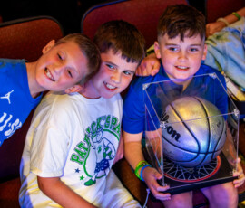 Three boys grin in the audience at North Shore Music Theatre, one holding a signed basketball in a display case, at A Night for Patrick