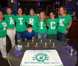 Volunteers in green shirts spelling PATRICK stand behind a sheet cake reading All In for Patrick at North Shore Music Theatre