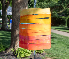 A hanging painted panel in layered sunset yellows, oranges and reds, slit into waves and mounted beside a tree on Beverly Common during Beverly's Garden Party