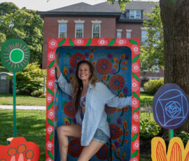 A visitor poses inside Ally Rowell's walk-in painted floral frame, surrounded by wooden flower cutouts staked into the grass at the Beverly Garden Party