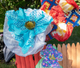 Oversized handmade flowers in turquoise lace, crocheted granny squares and beaded purple fabric displayed above a painted picket fence at the Beverly Garden Party