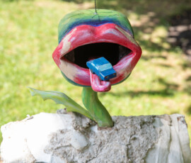 A sculpted flower shaped as an open mouth with pink lips, holding a small blue toy car on its tongue, mounted on a plaster base at the Beverly Garden Party
