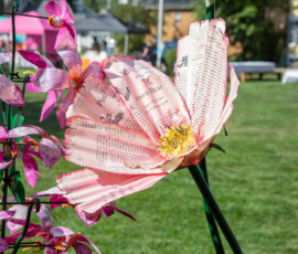 A large paper flower folded from printed book pages with dyed pink edges, staked beside magenta lilies at Beverly's Garden Party