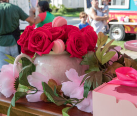 Red roses and pink macarons arranged in a ceramic urn beside a ribboned pastry box at a bakery table during the Beverly Garden Party