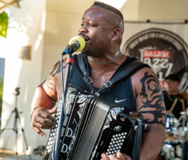 A musician with a mohawk and arm tattoos sings into a microphone while playing accordion on stage at the 2026 Salem Jazz and Soul Festival in Salem Willows. A drummer plays behind them, with a “Jazz Fest” banner partially visible in the background.