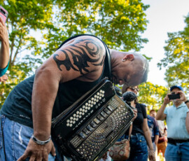 A man with a tribal tattoo on his arm leans forward playing an accordion outdoors at the 2026 Salem Jazz and Soul Festival in Salem Willows. Several people stand around him, some taking photos, with green trees and blue sky in the background.