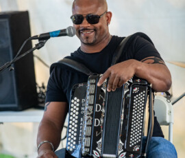 A man in sunglasses and a black t-shirt sits on a white chair, playing an accordion and smiling at a microphone during the 2026 Salem Jazz and Soul Festival at Salem Willows. He wears ripped blue jeans, with a large speaker and amplifier behind him.