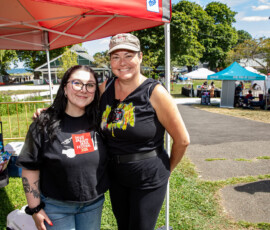 Two women stand smiling at an outdoor event under a red canopy at Salem Willows during the 2026 Salem Jazz and Soul Festival, with vendor tents, tables, and people enjoying a sunny day in the park.