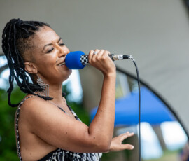 A woman with braided hair sings into a blue microphone at the 2026 Salem Jazz and Soul Festival in Salem Willows. Wearing a sleeveless patterned dress and large earrings, she performs expressively, trees and a blue tent blurred in the background.