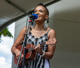 A person wearing geometric earrings and a patterned sleeveless top plays a ukulele and sings into a microphone onstage at the 2026 Salem Jazz and Soul Festival at Salem Willows.