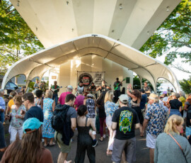 A festive crowd dances and enjoys live music at the Salem Jazz and Soul Festival 2026, held under a white canopy at Salem Willows. Sunlight filters through nearby trees as attendees watch the band perform on stage in this lively outdoor scene.