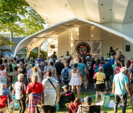 A large crowd gathers outdoors at Salem Willows in front of a white stage where a band performs for the 2026 Salem Jazz and Soul Festival. People are standing, sitting in chairs, and enjoying the music under a clear, sunny sky.