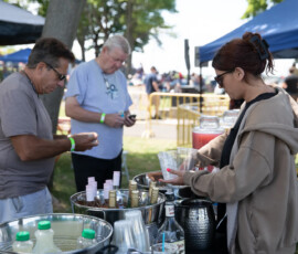 At the Salem Jazz and Soul Festival 2026 at Salem Willows, a woman in sunglasses prepares drinks at an outdoor table as two men stand nearby—one on his phone, the other holding a cup—amid drink dispensers, condiments, and plastic cups. Trees and tents set the scene.