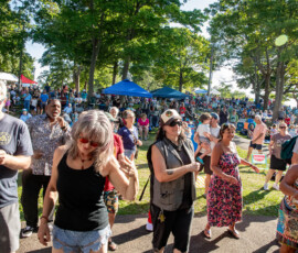 A large crowd gathers outdoors under trees at the Salem Willows Soul Festival. People of various ages stand, talk, and stroll around. Some wear hats and sunglasses. Blue tents are visible in the background, sunlight filters through the trees.