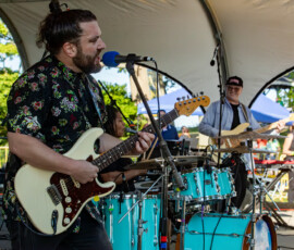 At the Salem Jazz and Soul Festival 2026, a band performs on an outdoor stage. The guitarist and lead singer stand at a microphone, while another plays electric bass. A turquoise drum set is in front; keyboard equipment and Salem Willows’ trees are seen beyond the stage.