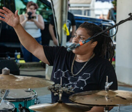 A drummer with shoulder-length hair sits at a drum set, raising one arm while speaking into a microphone. Wearing a black shirt with a chemical structure graphic, they perform at Salem Jazz in Salem Willows as a photographer captures the Soul Festival vibe.