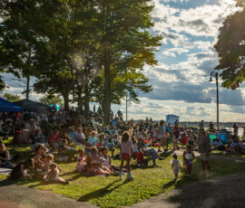 A large crowd, including families with children, gathers on the grass under trees at Salem Willows for the 2026 Salem Jazz and Soul Festival. The sun shines through the trees, and tents and equipment are visible under a partly cloudy sky.
