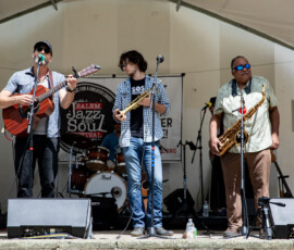 Three musicians perform on an outdoor stage at the Salem Jazz and Soul Festival 2026 in Salem Willows. One plays guitar and sings, another holds a trumpet, and the third holds a saxophone. Drums are visible behind them with a festival banner overhead.