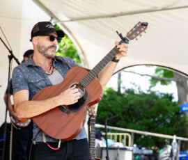 At the Salem Jazz and Soul Festival 2026, a man in sunglasses, a cap, and short sleeves strums an acoustic guitar onstage. Music equipment surrounds him as he performs with greenery and the vibrant energy of Salem Willows in the background.